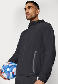 Giacca leggera nera con zip frontale, cappuccio e polsini elasticizzati. Presenta una tasca laterale con zip e logo "AEROREADY" in grigio riflettente.