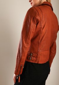 Kvinde iført en rustfarvet læderjakke med sidelukninger og en sort tekstureret nederdel, delvist vendt væk fra kameraet.