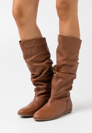 Bottes hautes marron en cuir avec un design décontracté, texture lisse, bout arrondi et semelle plate. Comprend une couverture de la cheville et des détails plissés.