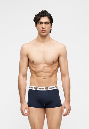 Homme musclé aux cheveux ondulés foncés portant un boxer noir Moschino avec une ceinture à ours en peluche et logo, debout devant un fond blanc uni.