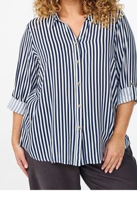 Chemise à rayures navy et blanche avec des manches courtes roulées, dotée de boutons dorés et d'une coupe décontractée.