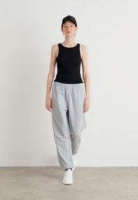 Zwarte tanktop, grijze sweatpants met elastische tailleband en witte sneakers. Model draagt een zwarte pet en staat in een minimalistische setting.