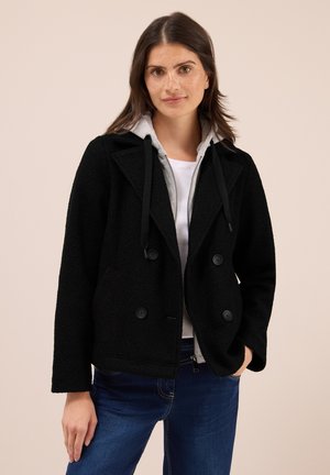 Veste en polaire noire avec une capuche grise, design à double boutonnage et boutons noirs. Portée sur une chemise blanche, associée à un jean bleu.