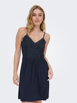 Ung kvinde iført en sort ærmeløs kjole med blonder bodice og spaghetti stropper, står foran en ensfarvet hvid baggrund.