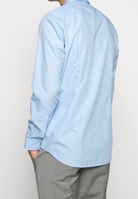 Lichtblauw langemouwen shirt met een zachte textuur, met knoopmanchetten en een ronde zoom, gezien vanaf de achterkant tegen een neutrale achtergrond.
