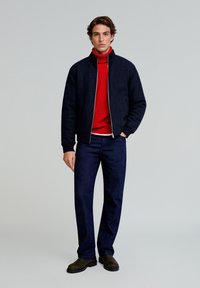 Chaqueta bomber azul marino con cremallera plateada, jersey de cuello alto rojo, jeans de mezclilla oscura y botas marrones. Diseño sencillo con un acabado texturizado.