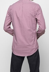 Červená a bílá kostkovaná košile s dlouhým rukávem, skladem v zádech a zakřiveným lemem, vyrobená z bavlněné látky. Temné džíny doplňují vzhled.