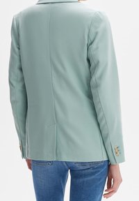 Mint green blazer, textured fabric, tailored fit, single vent at back, gold button accents on cuffs, paired with blue jeans.