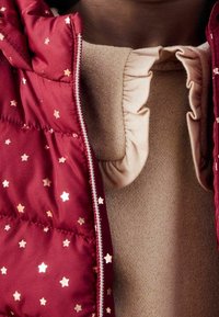 Chaleco rojo ligero con patrones de estrellas doradas, desabrochado para revelar una blusa beige con una superficie texturizada y detalles de cuello con volantes.