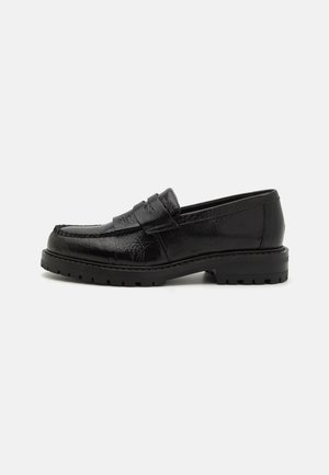Mocassins en cuir noir avec une finition texturée, présentant une sangle décorative, un bout arrondi et une semelle épaisse et robuste.