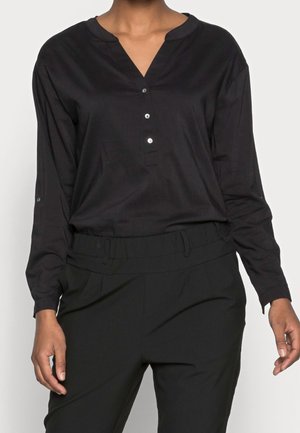 Personne portant une chemise noire à manches longues boutonnée rentrée dans un pantalon noir taille haute, debout devant un fond clair uni.