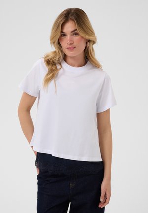 Jeune femme aux longs cheveux blonds portant un haut blanc uni à manches courtes et un pantalon foncé, debout devant un fond clair uni.