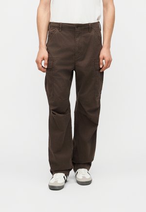 Persoană purtând pantaloni cargo largi maro închis cu buzunare laterale și adidași albi cu dungi negre, stând pe un fundal simplu.