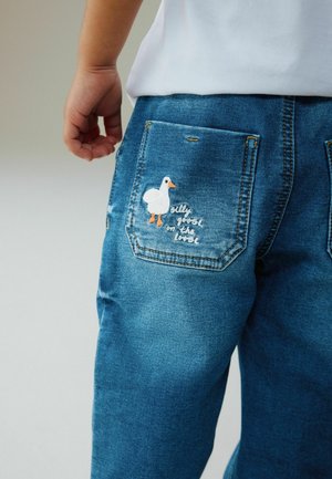 Denimjeans i mellemblå med en baglomme, der har en hvid og orange broderet and og teksten "silly goose on the loose".