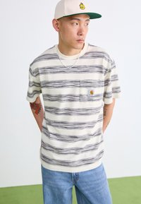Kremsstripet skjorte med kort erm, frontlomme og logo-patch. Kombinert med blå jeans. Har en lysfarget caps.