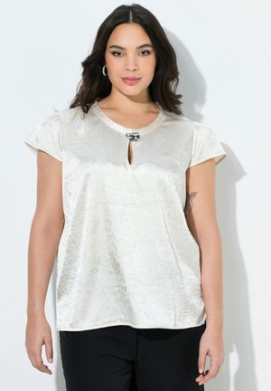 Vrouw met donker haar die een ivoorkleurige satijnen blouse met korte mouwen draagt, voorzien van een klein voorste keyhole en een decoratieve gesp, gecombineerd met zwarte broek.