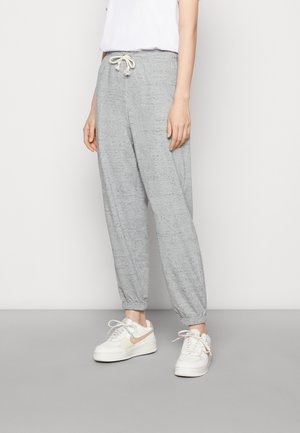 Persona che indossa pantaloni della tuta grigio chiaro con coulisse e caviglie risvoltate, e sneakers bianche con dettagli beige, in piedi su un pavimento grigio.