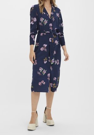Femme portant une robe portefeuille fleurie bleu marine à manches longues, une ceinture et des talons compensés crème sur un fond uni.