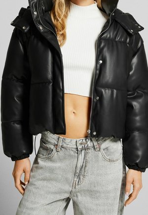 Femme portant une doudoune noire sur un pull court blanc à côtes et un jean taille haute gris clair, debout les bras détendus.