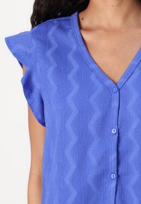 Maglietta blu con colletto a V e maniche corte. Il tessuto textured presenta un motivo a zigzag, con piccoli bottoni blu come accento.