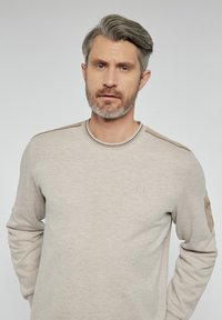Jasnobeżowy sweatshirt z okrągłym dekoltem, z fakturowanym szwem i zamszowymi łatami na rękawach. Dyskretne logo na klatce piersiowej. Gładki materiał.