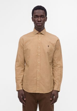 Homme portant une chemise à carreaux beige et marron avec des boutons et un petit logo brodé sur la poitrine, debout devant un fond blanc uni.