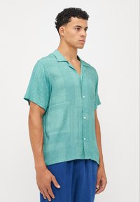 Camisa de manga curta em azul-turquesa claro com um corte descontraído, apresentando um padrão subtil e fecho frontal com botões. Tecido suave com uma textura lisa.