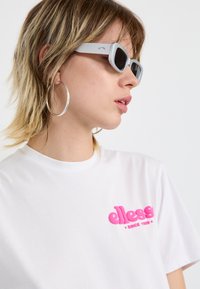 Hvit t-skjorte med rosa logo brodert på venstre bryst, kombinert med lysegrå rektangulære solbriller og store sølv hoop øredobber.