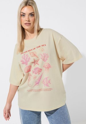 Donna bionda che indossa una t-shirt beige oversize con creature marine rosa e testo rosso "Tesori del Mare", abbinata a jeans blu.