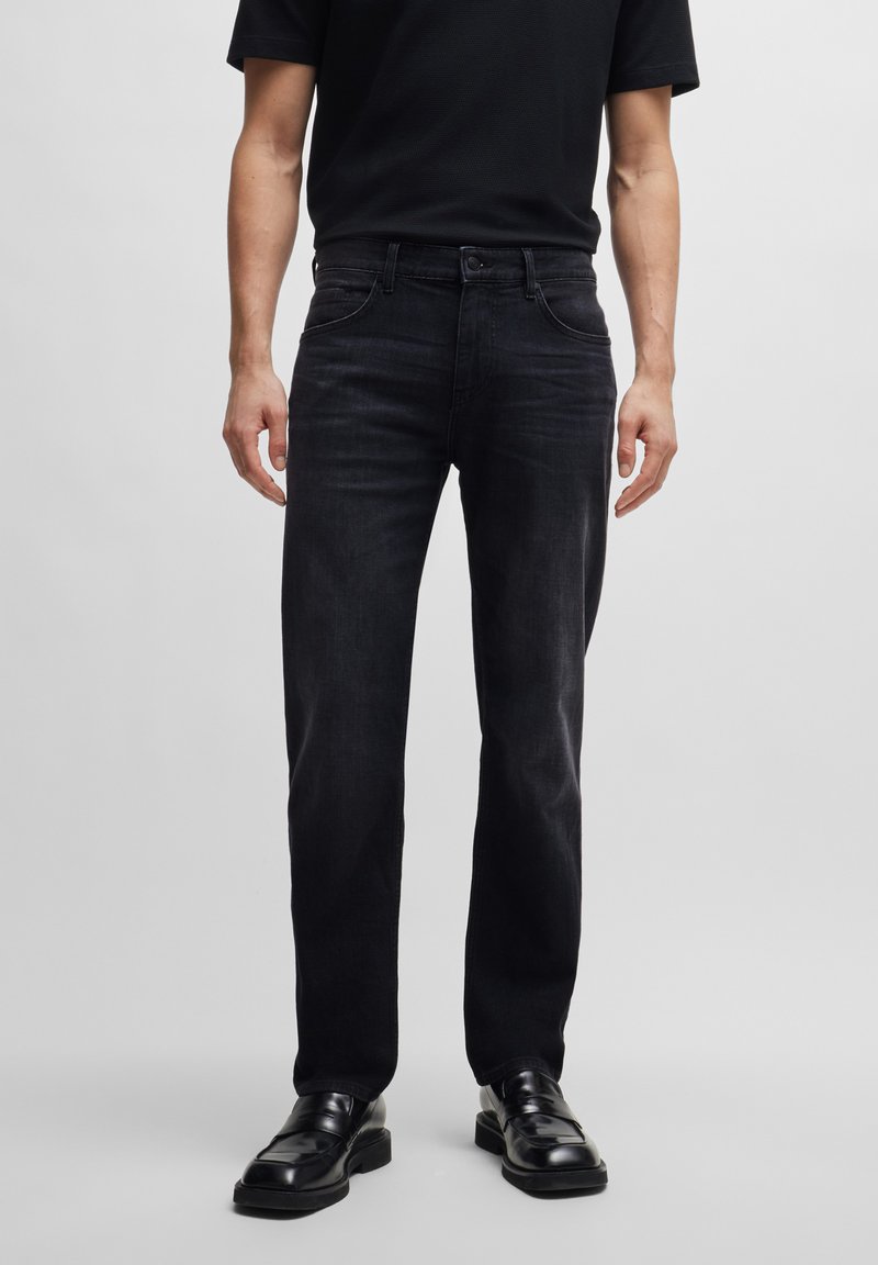 Schwarze Denim-Jeans mit geradem Schnitt, subtiler Ausbrenner-Textur, fünf Taschen und Knopfverschluss. Kombiniert mit schwarzen Schuhen und einem schwarzen Shirt.