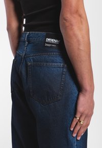 Tmavě modré denimové džíny s vysokým pasem, obsahující dvě zadní kapsy a štítek s logem na pásku. Hladká textura s minimálním vyblednutím.