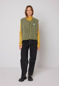 Groene fleece vest met een ronde hals, knopen aan de voorkant en een blauwe logopatch, gedragen over een mosterdkleurige coltrui en zwarte jeans.