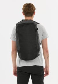 Musta reppu, jossa on teksturoitu pinta, rullatoppinen muotoilu ja säädettävät hihnat, esitettynä henkilön päällä, joka on pukeutunut harmaaseen paita ja tummiin shortseihin.