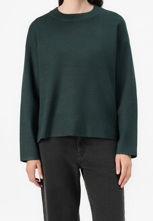 Pull en maille vert foncé à col rond manches longues porté avec un jean noir taille haute sur une personne debout.