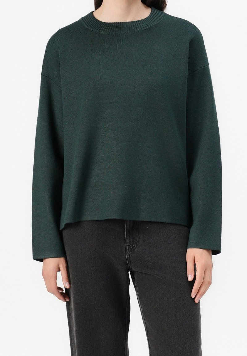 Pull en maille vert foncé à col rond manches longues porté avec un jean noir taille haute sur une personne debout.