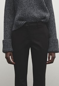 Pull gris en maille côtelée avec larges poignets, associé à un pantalon noir ajusté et élégant, doté d'une fermeture à boutons et d'un design épuré.