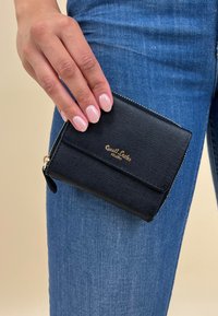 Schwarze, strukturierte Geldbörse aus Leder mit einem goldenen Logo und Reißverschluss, gehalten gegen blaue Jeans.