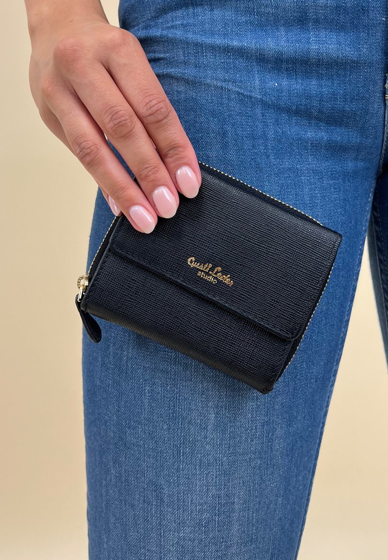 Cartera negra texturizada de cuero, con un logo dorado y cremallera, sostenida contra unos jeans de mezclilla azul.