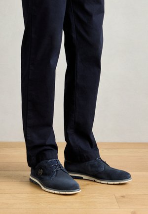 Zapatos de cordones azul marino con parte superior texturizada, suela blanca contrastante y punta redonda. Llevados con pantalones de tela oscura sobre un suelo de madera.