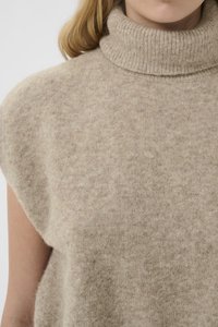 Frau trägt einen beigen, ärmellosen, hochgeschlossenen Strickpullover mit weicher, flauschiger Textur vor einem einfarbigen Hintergrund.