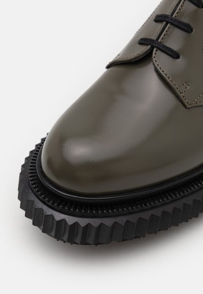 Olive green polished leather shoe with a thick black rubber sole featuring a serrated edge. Black laces and subtle stitching detail.