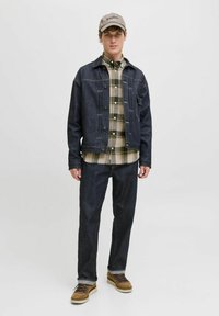 Chaqueta de mezclilla oscura con costuras en contraste, camisa de cuadros y jeans oscuros. Zapatos marrones completan el conjunto. La gorra tiene un logo bordado.