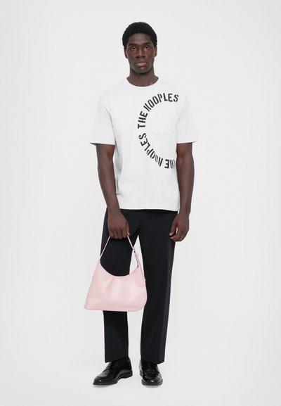 Biała koszulka z krótkim rękawem z czarnym nadrukiem, kontrastowa kieszeń, czarne spodnie, czarne buty, trzymająca miękką różową torbę na zamek.