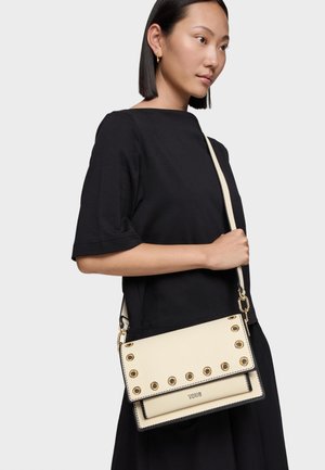 Mujer con cabello liso y negro, vestida con blusa y falda negras, sosteniendo un bolso cruzado color crema con detalles de ojales dorados.