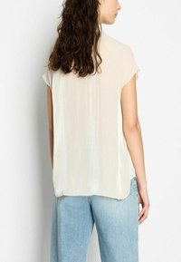 Top corto a maniche corte color beige chiaro con una superficie testurizzata e lucida, abbinato a jeans blu chiaro. Il tessuto morbido e fluido si drappeggia con eleganza.