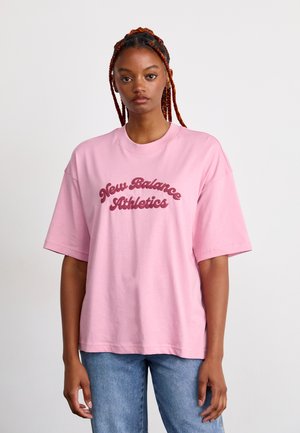 Young woman with braided hair wearing a loose pink "New Balance Athletics" t-shirt and blue jeans against a plain light background.