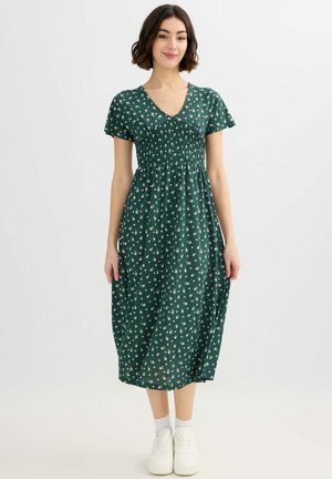 Joven mujer de pie, vistiendo un vestido midi verde oscuro de manga corta y escote en V con patrones florales blancos y zapatillas blancas.