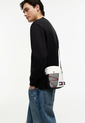 Junger Mann in schwarzem Sweatshirt und blauen Jeans, der eine weiße Umhängetasche mit Netzfach und roten Bungee-Seil-Details trägt.