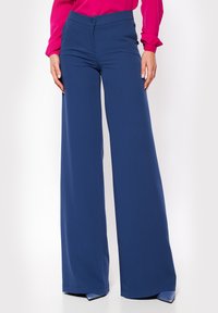 Kobieta w granatowych, szerokich, wysokich spodniach oraz jaskraworóżowej bluzce z długim rękawem, stoi z rękami swobodnie opuszczonymi wzdłuż ciała.