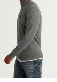 Hellgrauer Strickpullover mit ribbeligem Kragen und Bündchen, versehen mit einem kleinen schwarzen Logopatch auf der Brust. Getragen über einem weißen Hemd.